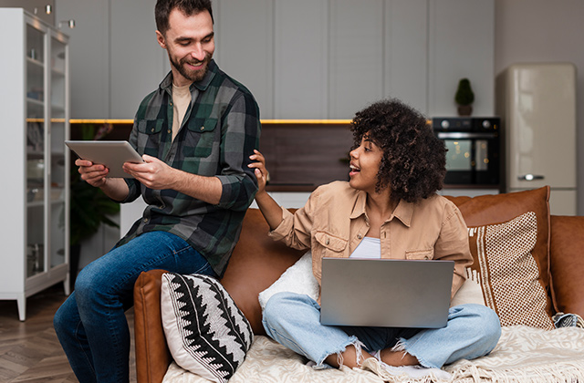 Black Friday Online Shopping is the perfect way for busy people to save money just before the holidays. A young couple is sitting on a couch comparing online discounts on their laptop and tablet.