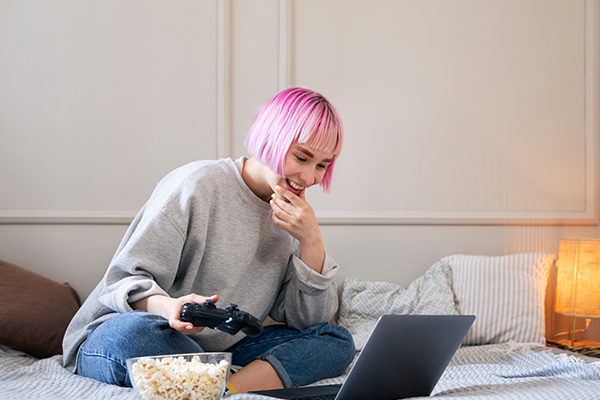 Eine junge Frau hält einen Controller und freut sich über ein Edutainment Game auf ihrem Laptop, das sie auf ihrem Bett sitzend spielt.