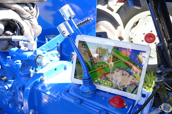 Zwischen den Schalthebeln eines blauen Oldtimer Traktors klemmt ein Tablet mit der Ansicht des Wirtshauses in My Free Farm 2, dem kostenlosen Farmspiel von upjers.
